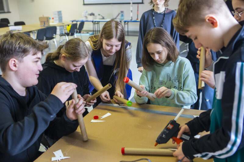 Grupa dzieci siedzi przy stole i buduje proste modele szybowców podczas warsztatów w Kopalni Ignacy