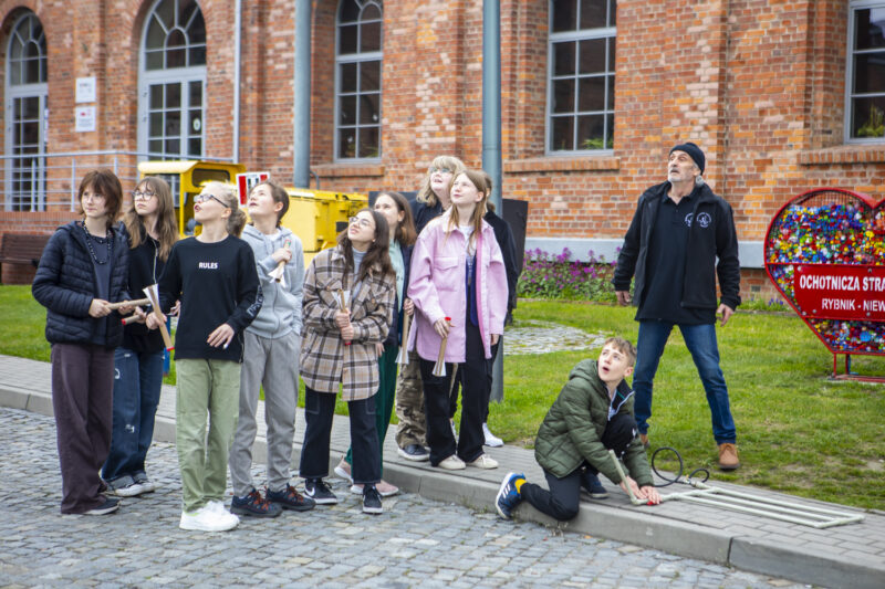 Dzieci na warsztatach naukowych w Kopalni Ignacy stoją przed budynkiem kopalni i patrzą w niebo.